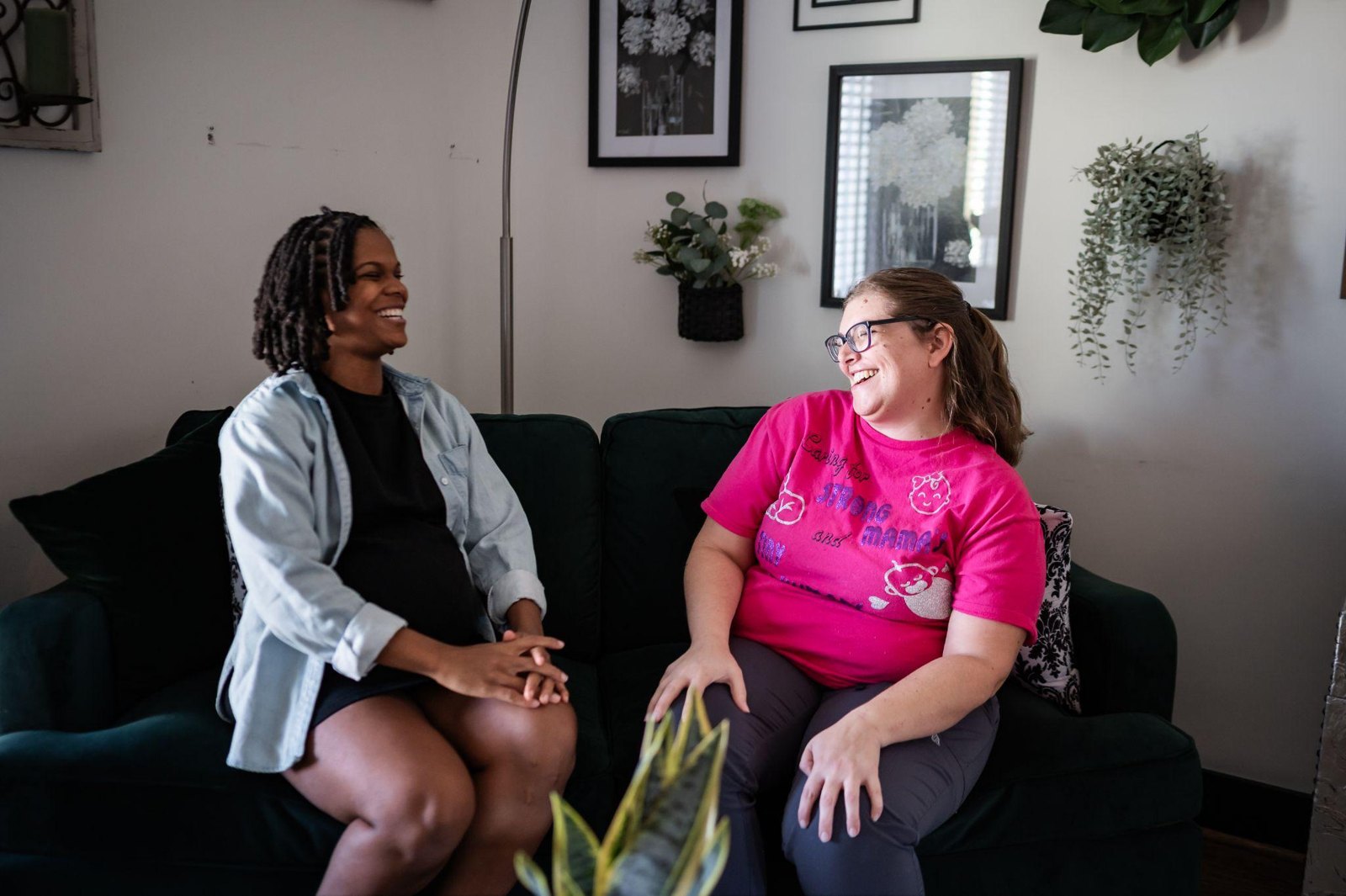 Two women sitting on a couch, one pregnant and one wearing a pink shirt, smiling and talking